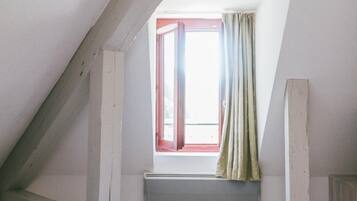 Un dormitori amb sostre inclinat, una finestra amb cortines, un radiador i un llit amb edredó blanc i rosa.