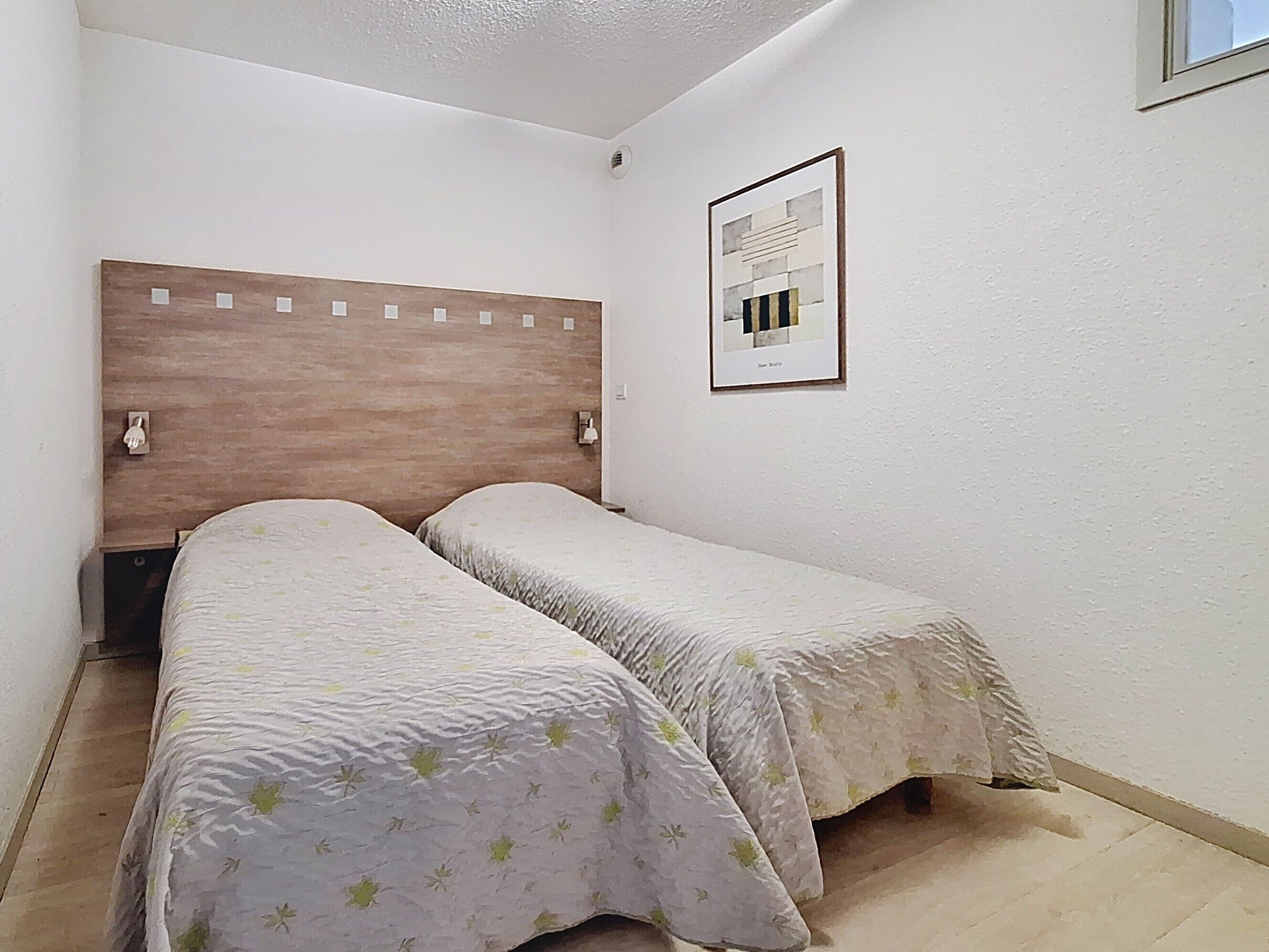 A small, single-room hotel room with a wooden headboard, two beds with floral bedding, a framed picture on the wall, and a window.