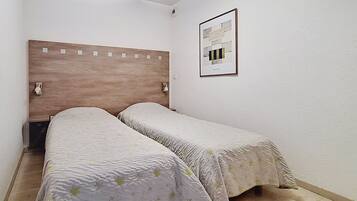A small, single-room hotel room with a wooden headboard, two beds with floral bedding, a framed picture on the wall, and a window.