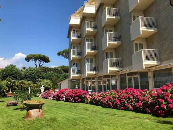 A multi-story hotel with balconies, a well-maintained garden with pink flowers, and a clear blue sky. - Hotel Atlantico (Forte dei Marmi)