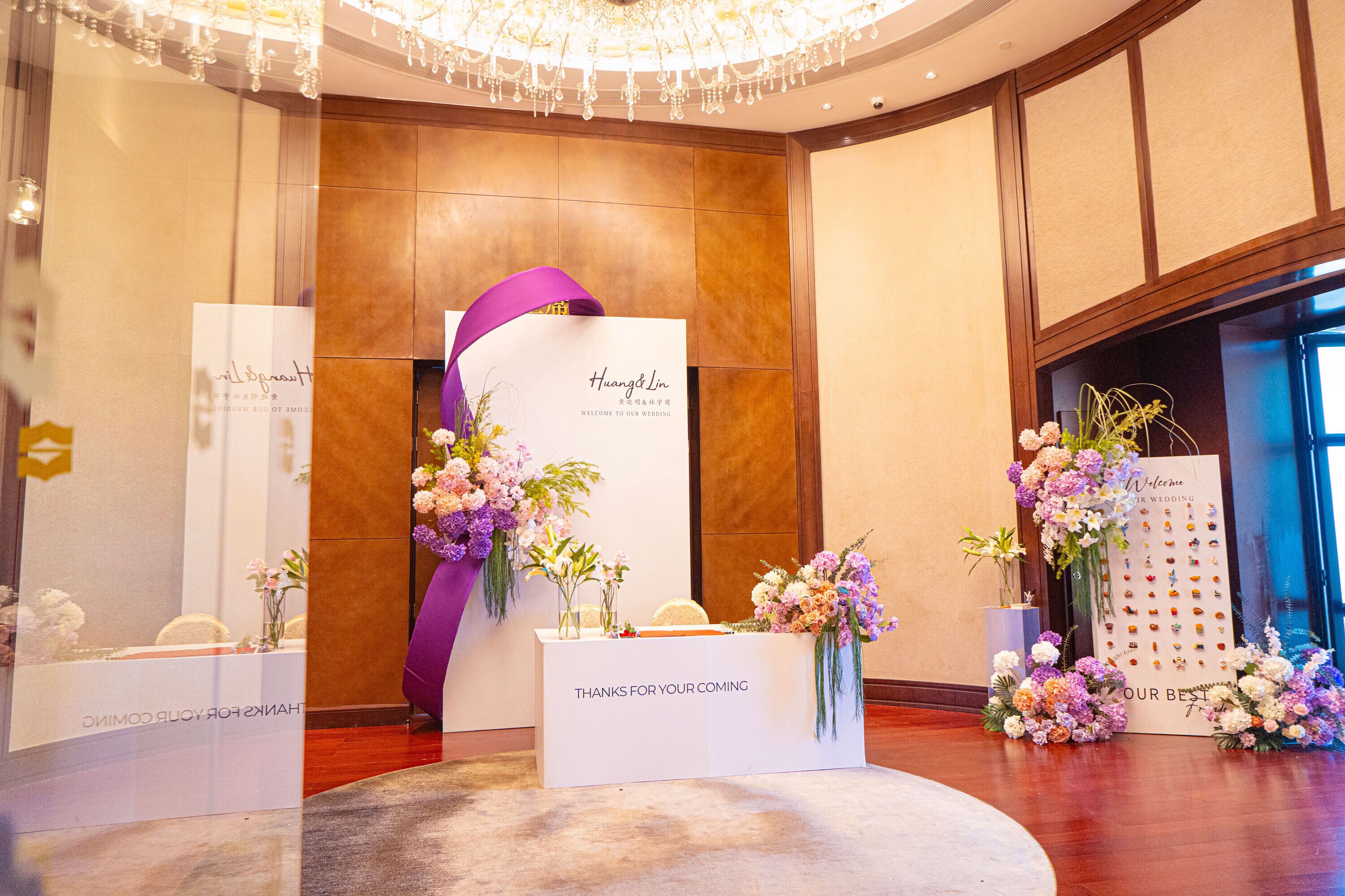 Zona de recepción con mostrador blanco, una cinta morada y arreglos florales.