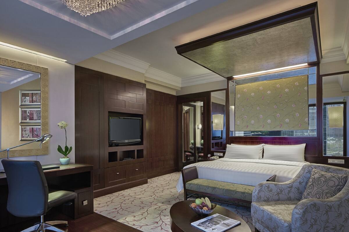Habitación de hotel con una cama grande, un escritorio con computadora, televisor, una silla y una planta decorativa.