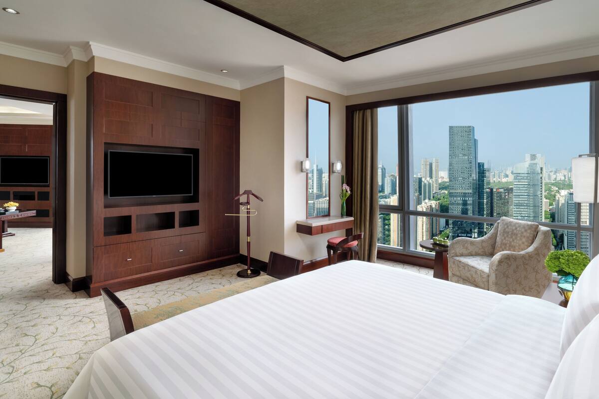 Habitación de hotel con una cama grande, televisor montado en la pared, vistas a la ciudad y un área de descanso con una silla y una mesa.