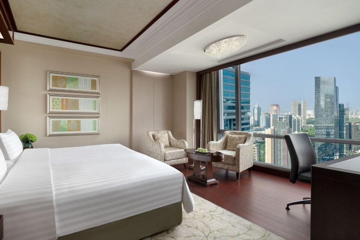 A hotel room with a large bed, two armchairs, a desk, and a view of a city skyline.