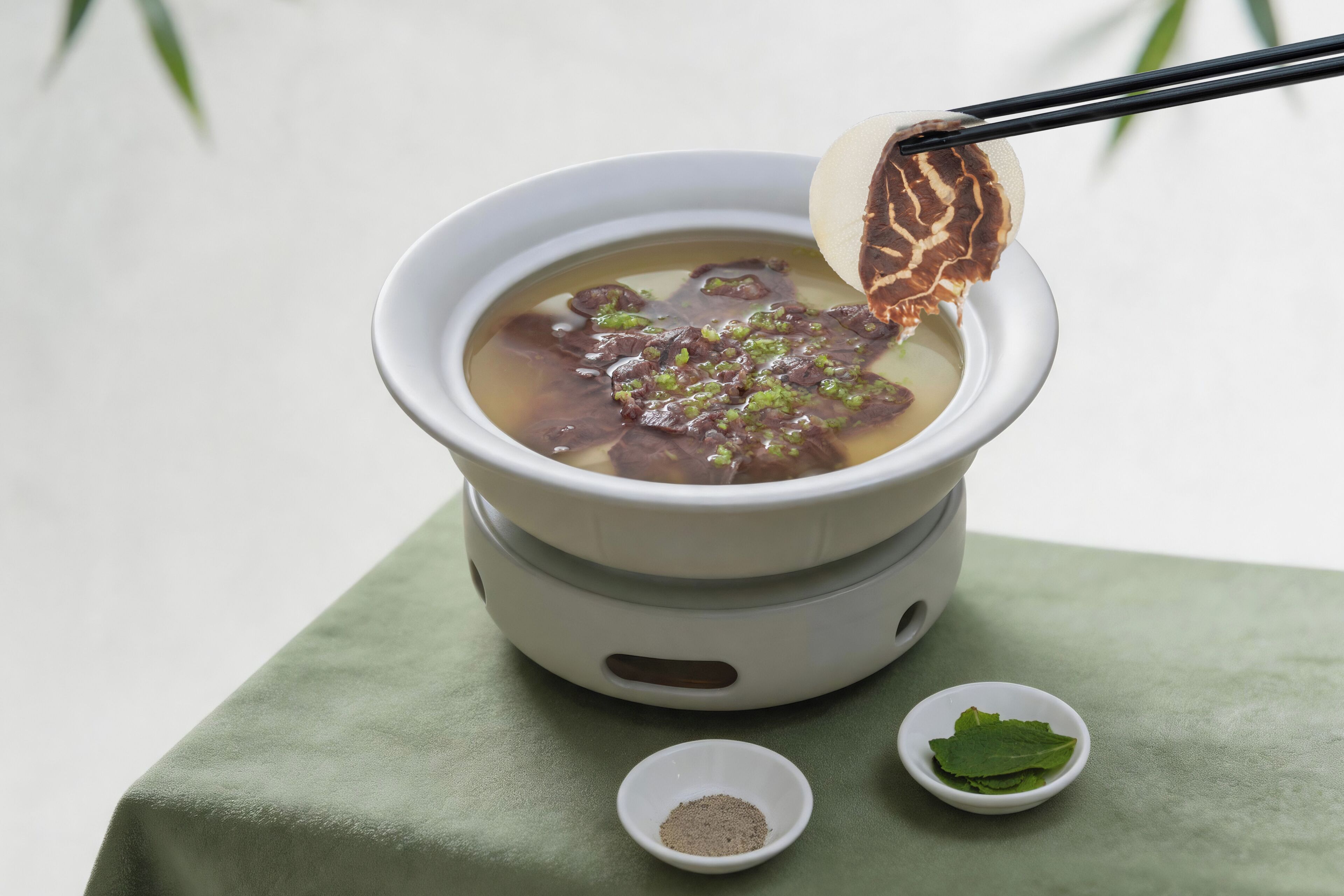Un bol de soupe avec des baguettes, un petit bol de feuilles vertes et un petit bol d’épices.