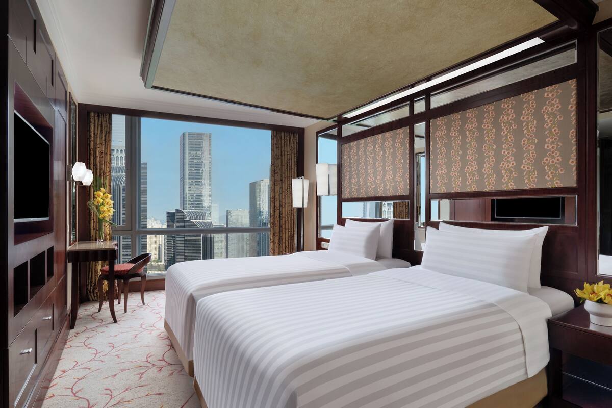 Habitación de hotel con dos camas, un escritorio con silla, televisor y un ventanal con vistas a la ciudad.