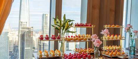 A table with tiered dessert displays and flowers, set against a cityscape view through large windows.