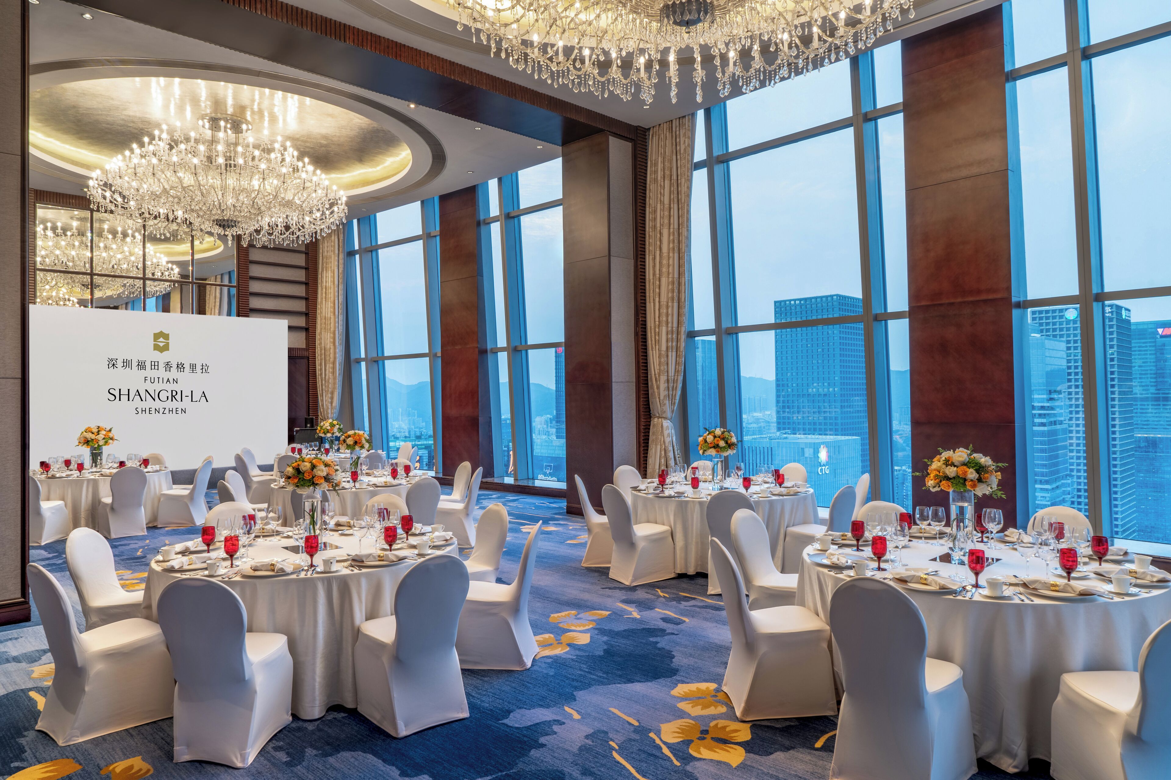 A banquet hall with round tables, white chairs, chandeliers, and a city view.