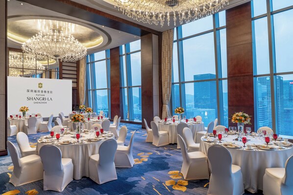 Un salón de banquetes con mesas redondas, sillas blancas, candelabros y vistas a la ciudad.