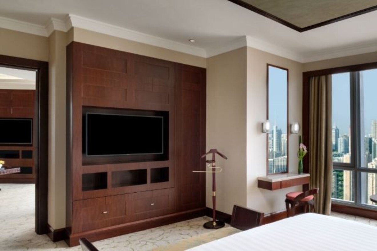 A hotel room with a large flat-screen TV mounted on a wooden panel wall, a bed with a white comforter, a desk with a chair, and a large window offering a city view.