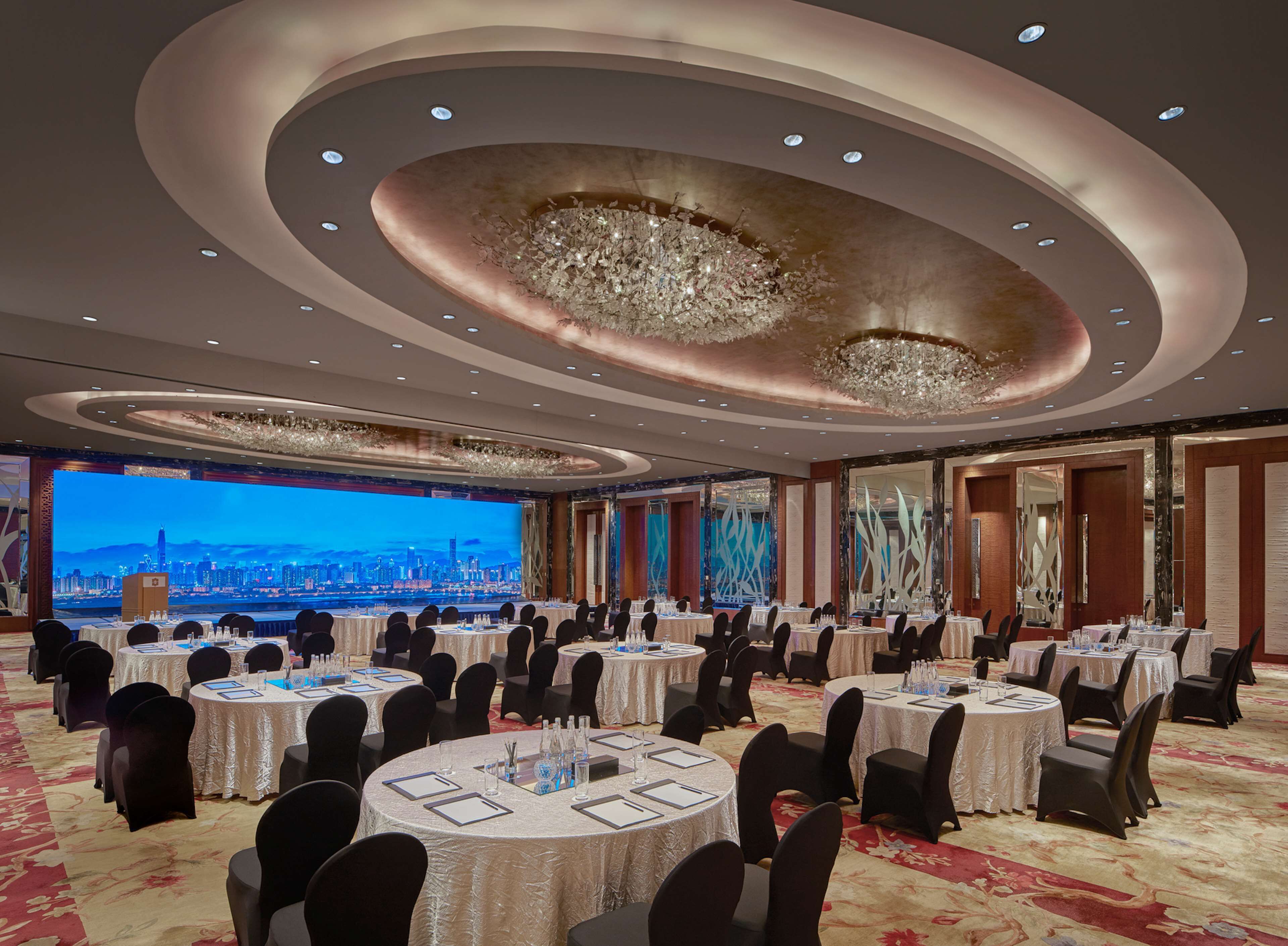 Un salón de banquetes con varias mesas redondas, sillas negras y una pantalla grande que muestra un paisaje urbano.