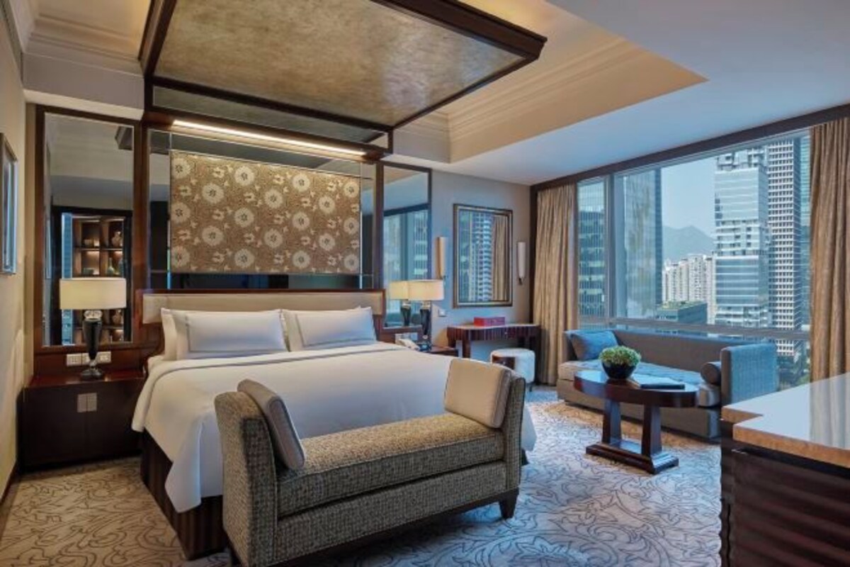 A hotel room with a large bed, a sitting area with a sofa and armchair, a small table, and a large window with a city view.