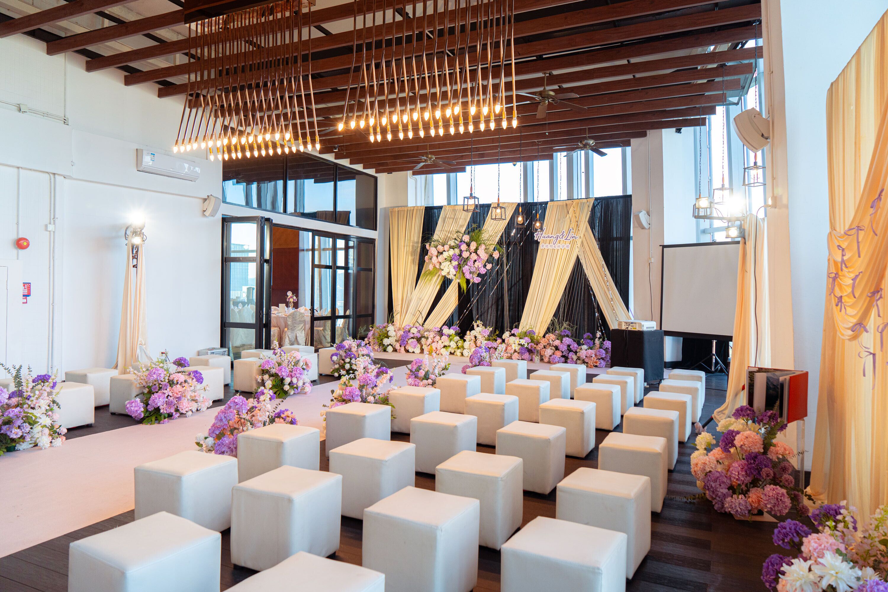A spacious hall with white cube seating arranged in rows, a stage with floral decorations, and a modern ceiling with hanging lights.