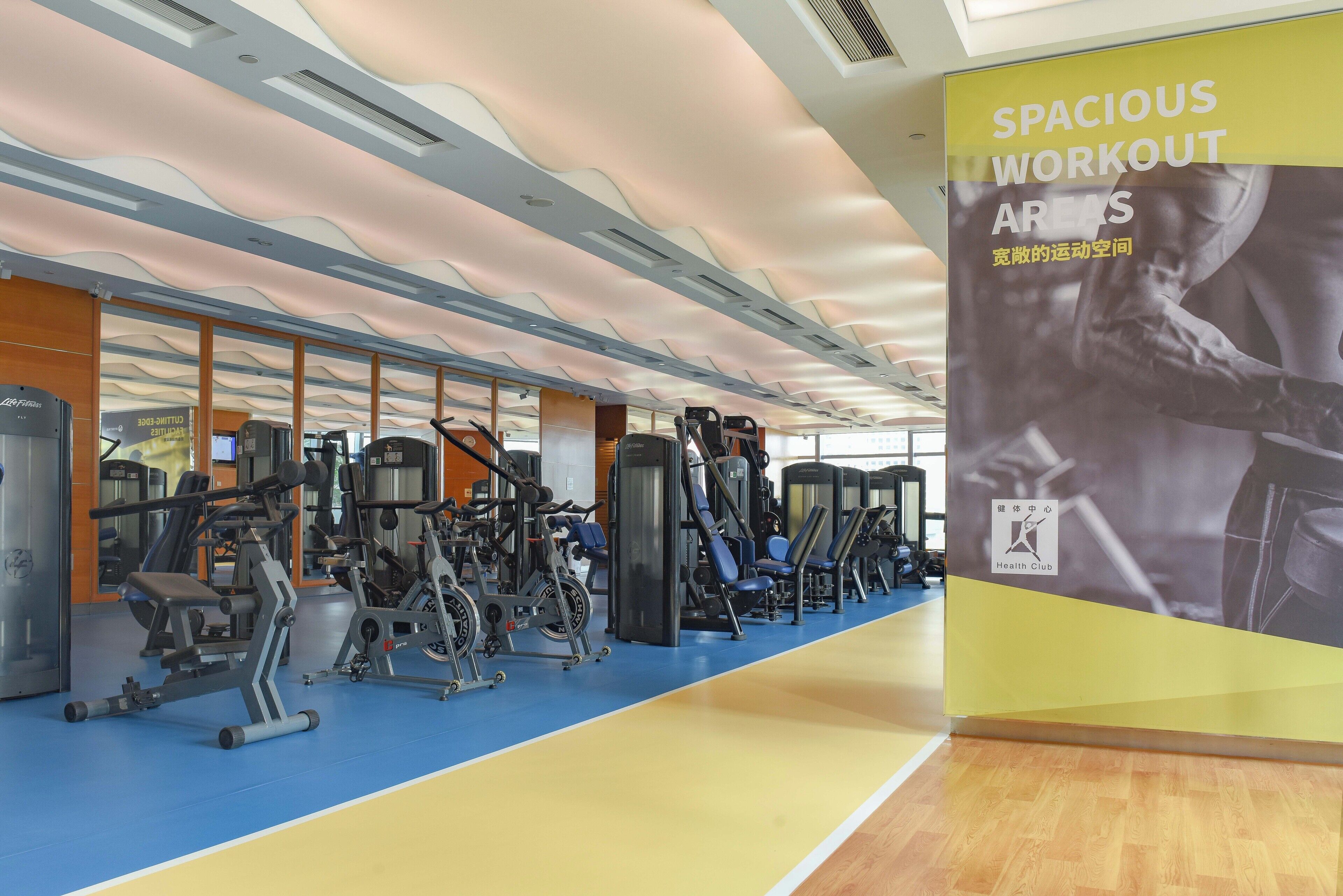 Un gimnasio con diversas máquinas de ejercicio, suelo azul y amarillo, y un cartel que anuncia amplias zonas de entrenamiento.