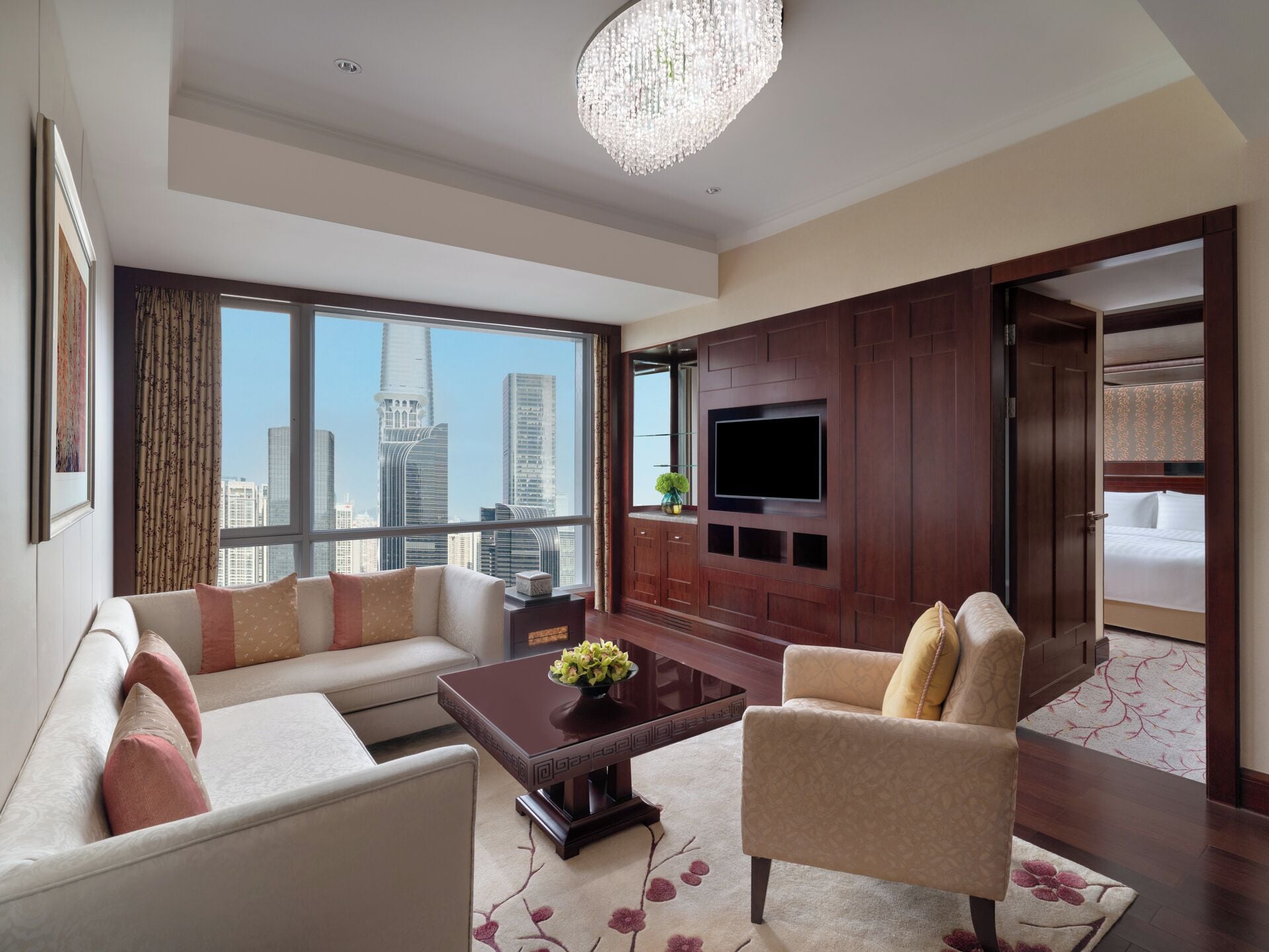 A modern hotel room with a large window offering a city view, a flat-screen TV mounted on a wooden panel, a sofa, a chair, and a coffee table with a floral arrangement.