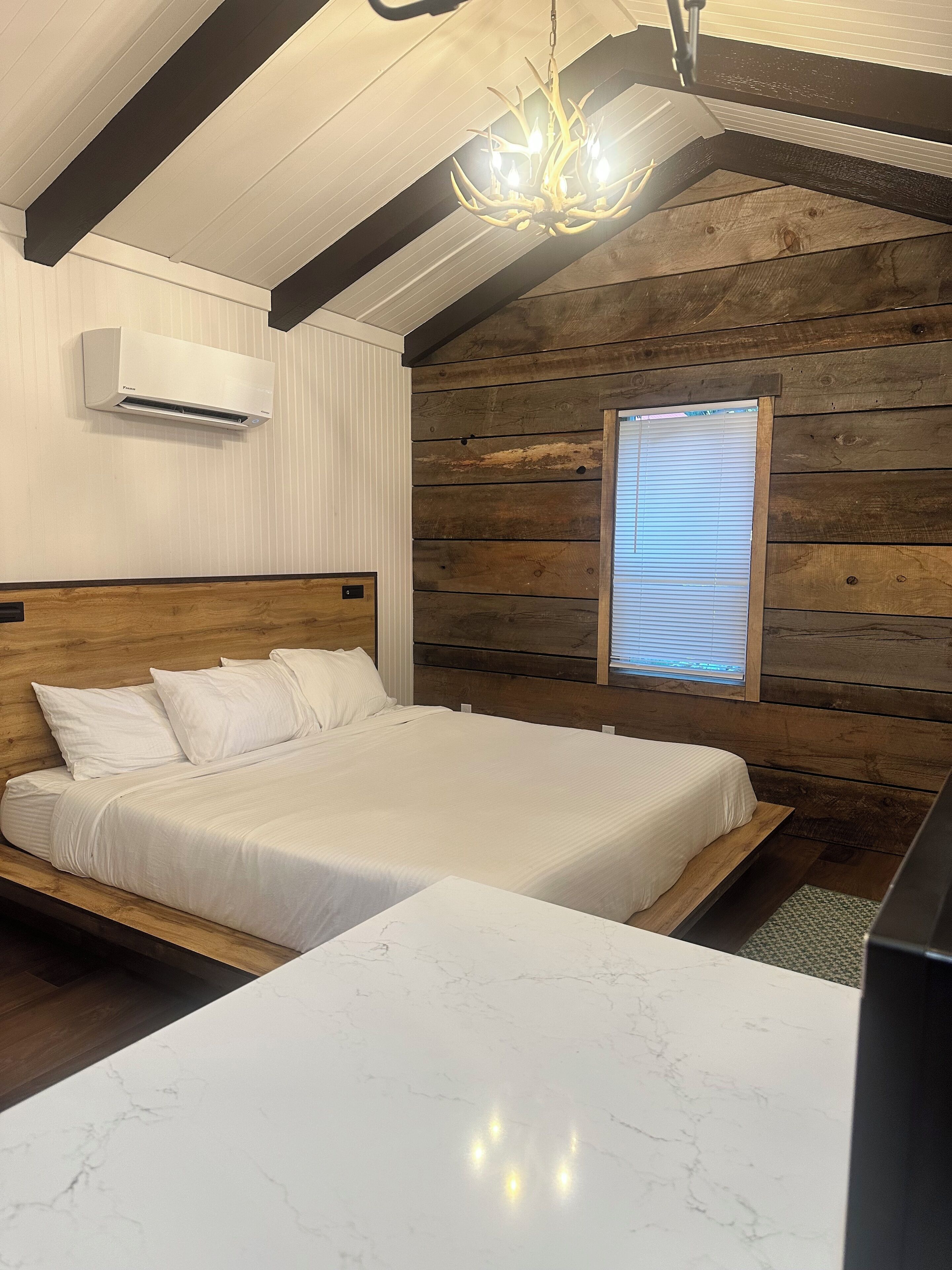 Un dormitorio con cabecera de madera, cama con ropa blanca, mesita de noche de mármol, candelabro y ventana con persianas.