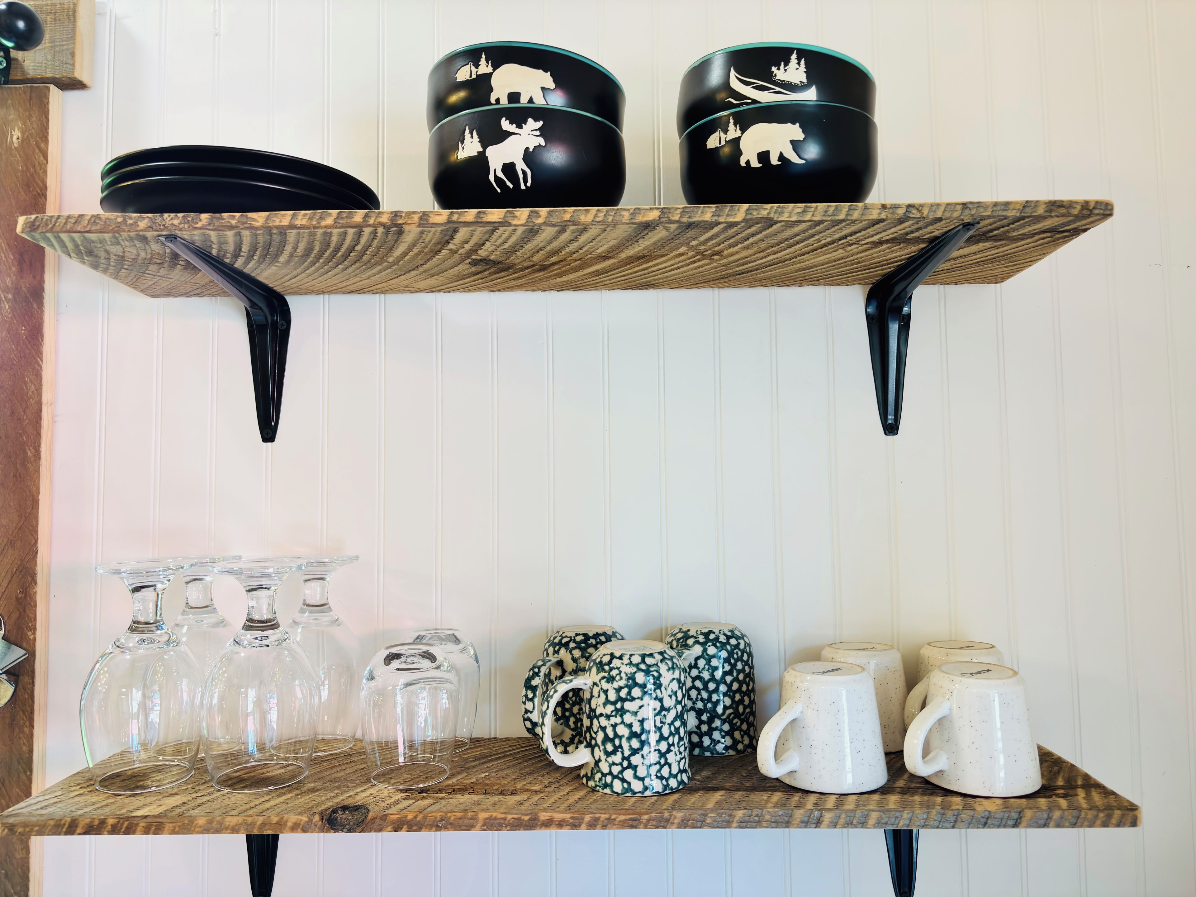Una estantería de madera con tazas blanco y negro, copas de vino transparentes y una taza estampada.