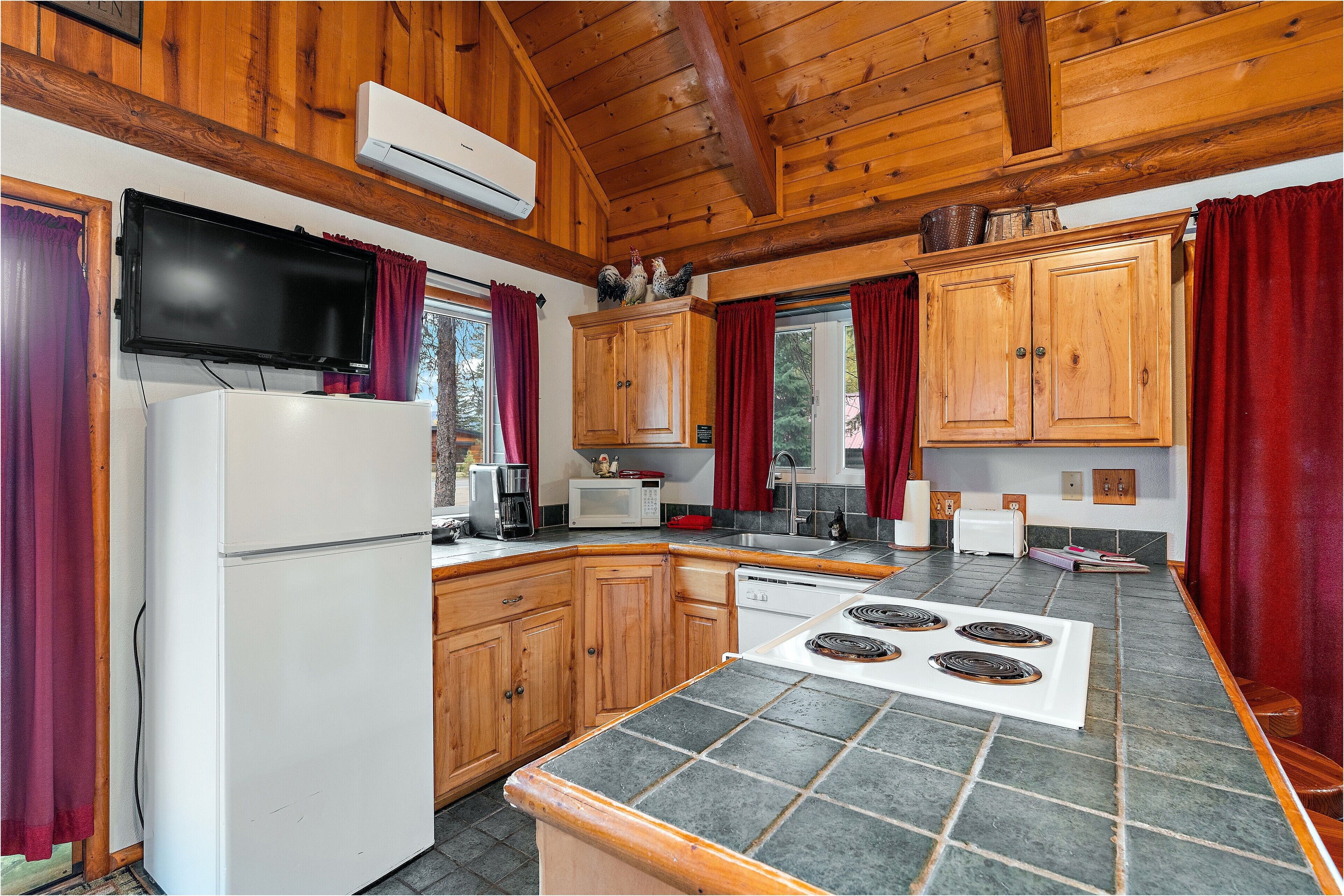 Una cocina con armarios de madera, un refrigerador blanco, un microondas, una estufa y cortinas rojas.
