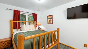 A bedroom with a wooden bed, a window with red curtains, a mounted TV, and a framed picture on the wall.