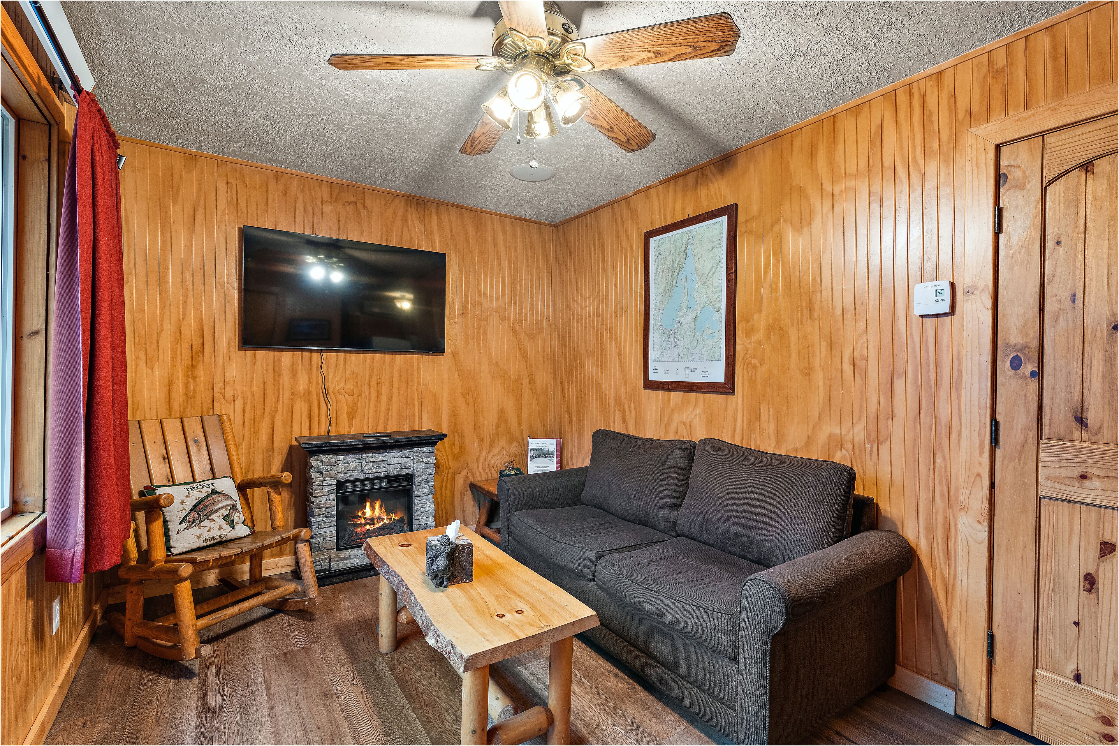 A cozy room with wooden walls, a fireplace, a sofa, a rocking chair, and a ceiling fan.