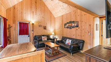 Un acogedor interior de cabaña con paredes de madera, un sofá de cuero negro, una mesa de centro de madera y un televisor de pantalla plana.