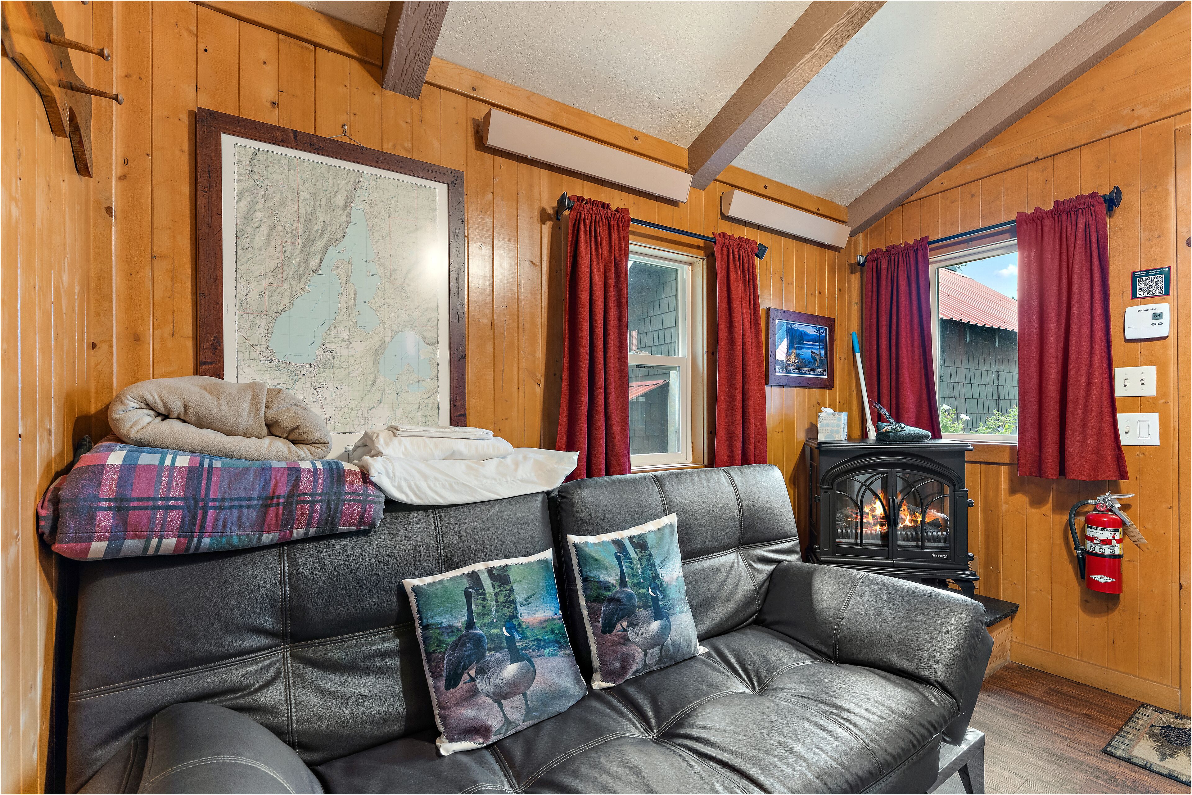 Una habitación acogedora con un sofá de cuero, dos almohadones decorativos, un mapa en la pared, una chimenea y cortinas rojas.