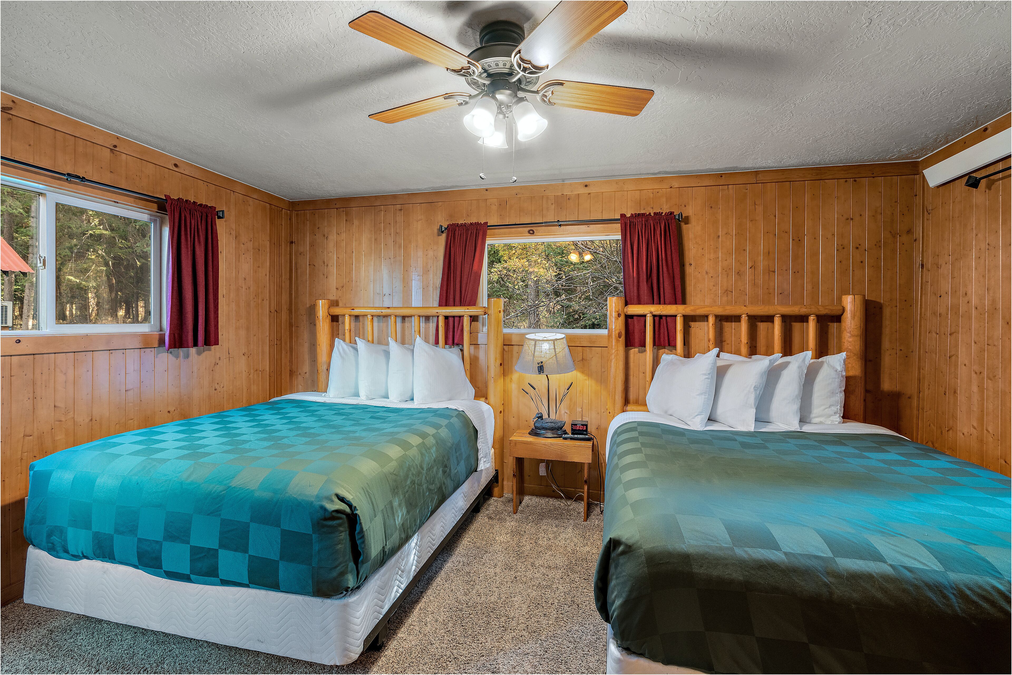 A room with two beds, wooden walls, a ceiling fan, and a window with curtains.