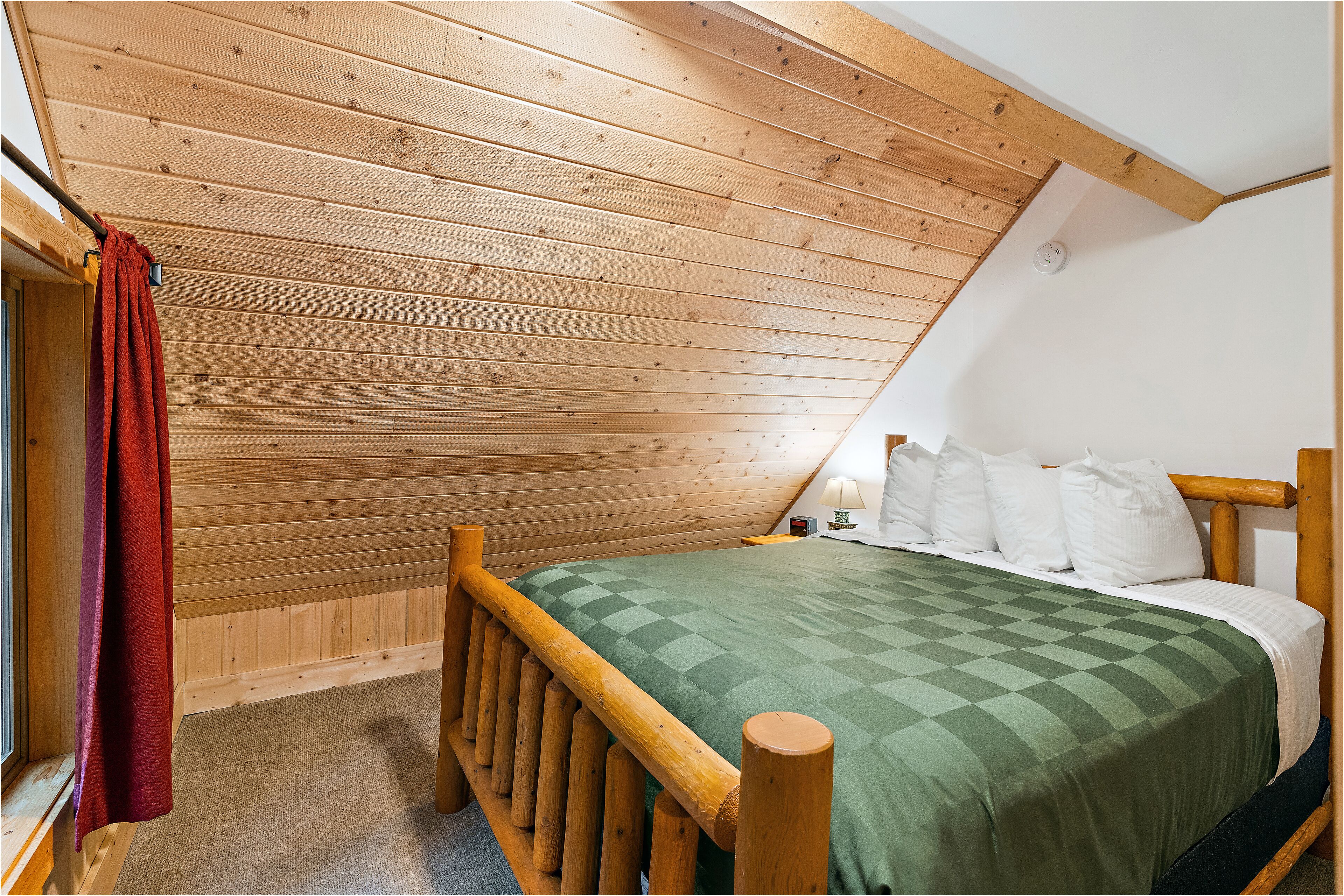 A bedroom with a sloped ceiling, wooden walls, a bed with a green checkered bedspread, white pillows, and a wooden headboard.