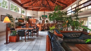 Un hall d’entrée spacieux avec un plafond haut, des poutres en bois et de grandes fenêtres. On y trouve plusieurs coins salon, dont un canapé et des fauteuils, ainsi qu’un bar avec un comptoir et des tabourets.