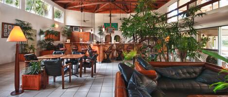 Un hall d’entrée spacieux avec un plafond haut, des poutres en bois et de grandes fenêtres. On y trouve plusieurs coins salon, dont un canapé et des fauteuils, ainsi qu’un bar avec un comptoir et des tabourets.