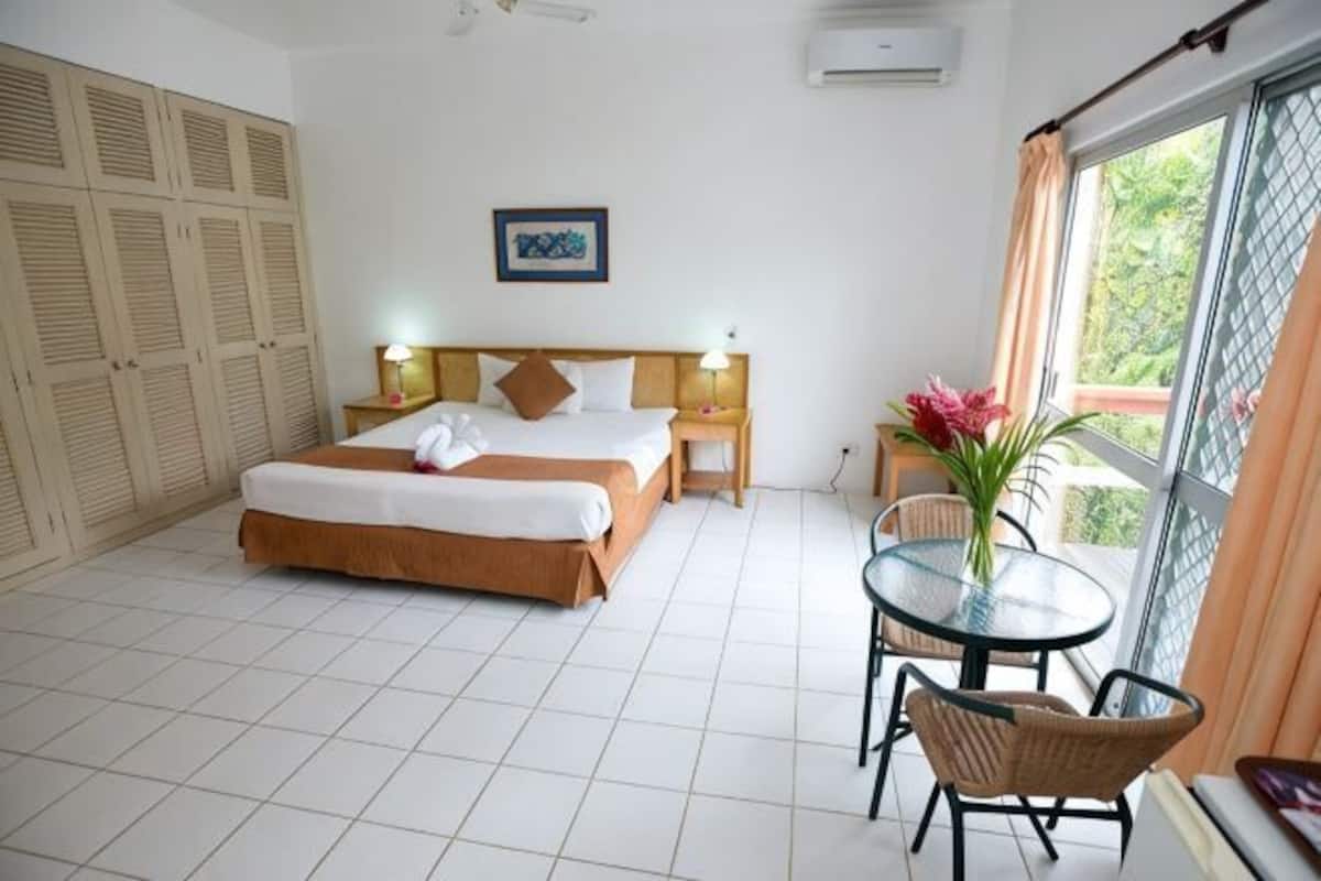 A hotel room with a bed, bedside tables, a small table with chairs, a window with curtains, and a view of greenery outside.