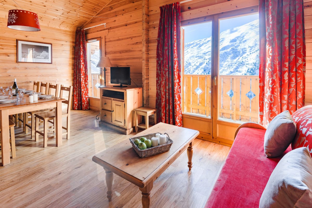 A cozy cabin with a wooden interior, a dining area, a television, and a view of a snowy mountain.