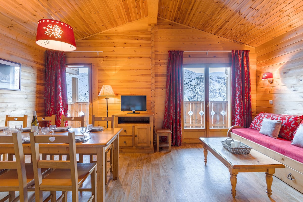 A wooden cabin interior with a dining table, chairs, a sofa, a television, and a window with curtains.