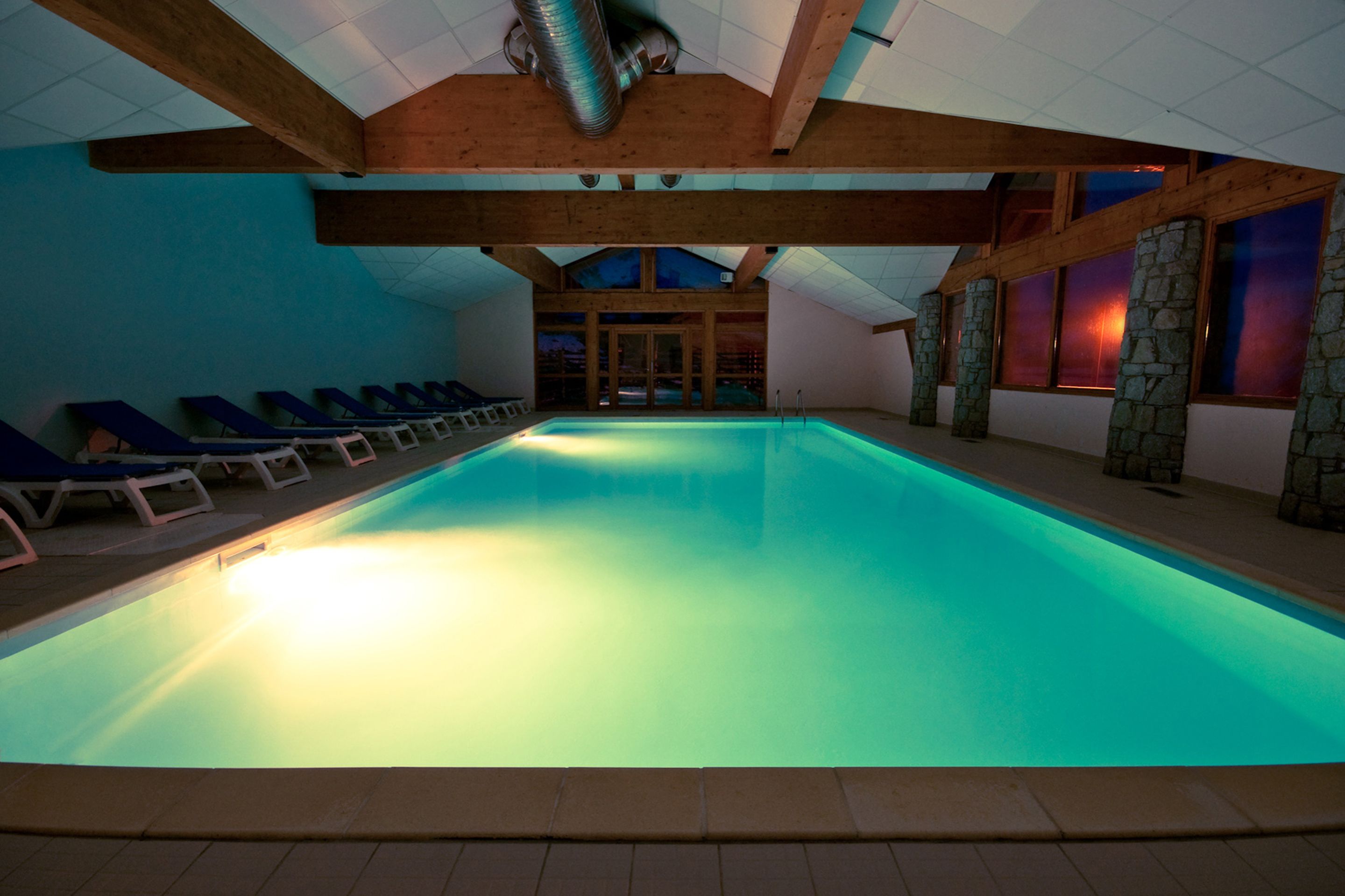 Een binnenzwembad met blauw verlichte sfeerverlichting, houten balken, stenen zuilen en loungestoelen.