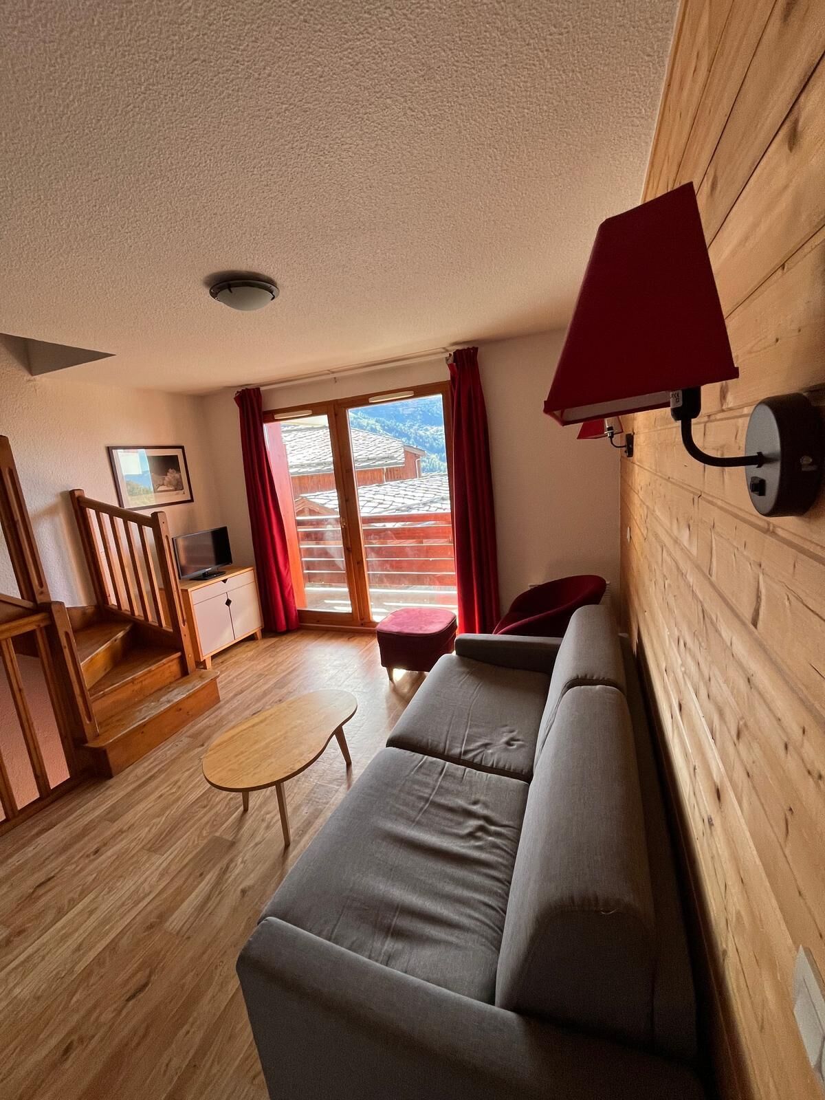 Una sala de estar acogedora con un sofá, una mesita y un televisor. Se ve una escalera que lleva a otra habitación, y una ventana con cortinas.