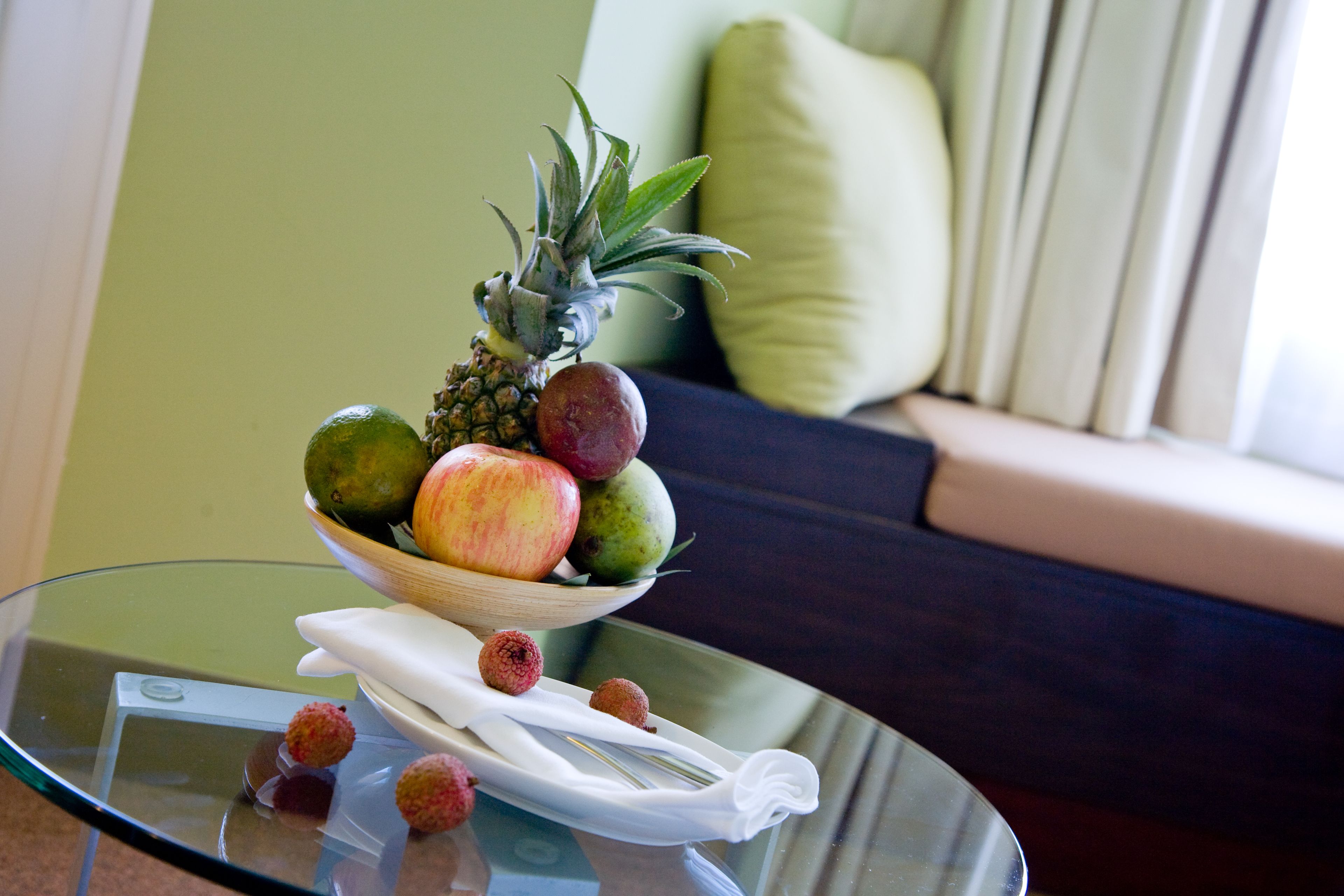 Una mesa de vidrio con un frutero que contiene piña, manzana y lima, además de un paño blanco.