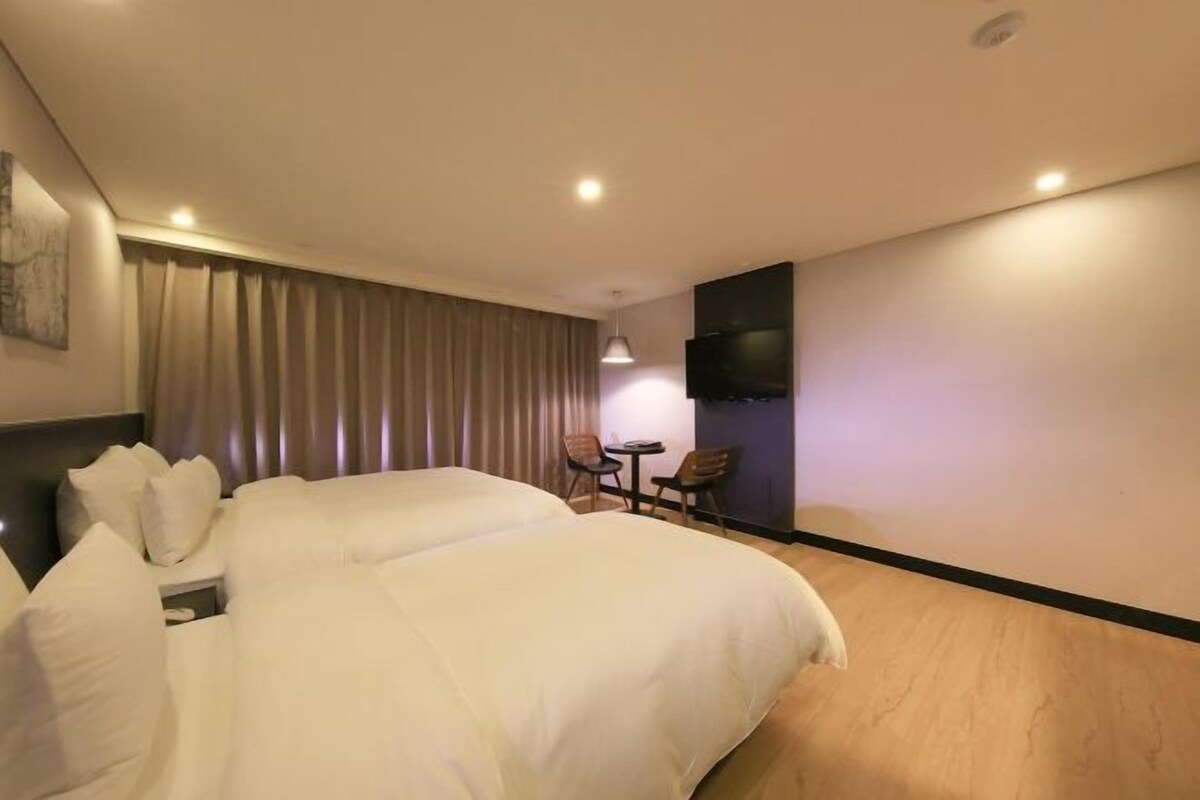 Ein Hotelzimmer mit einem großen Bett, einem Schreibtisch mit zwei Stühlen, einem Flachbildfernseher und einem an der Wand hängenden Kunstwerk.