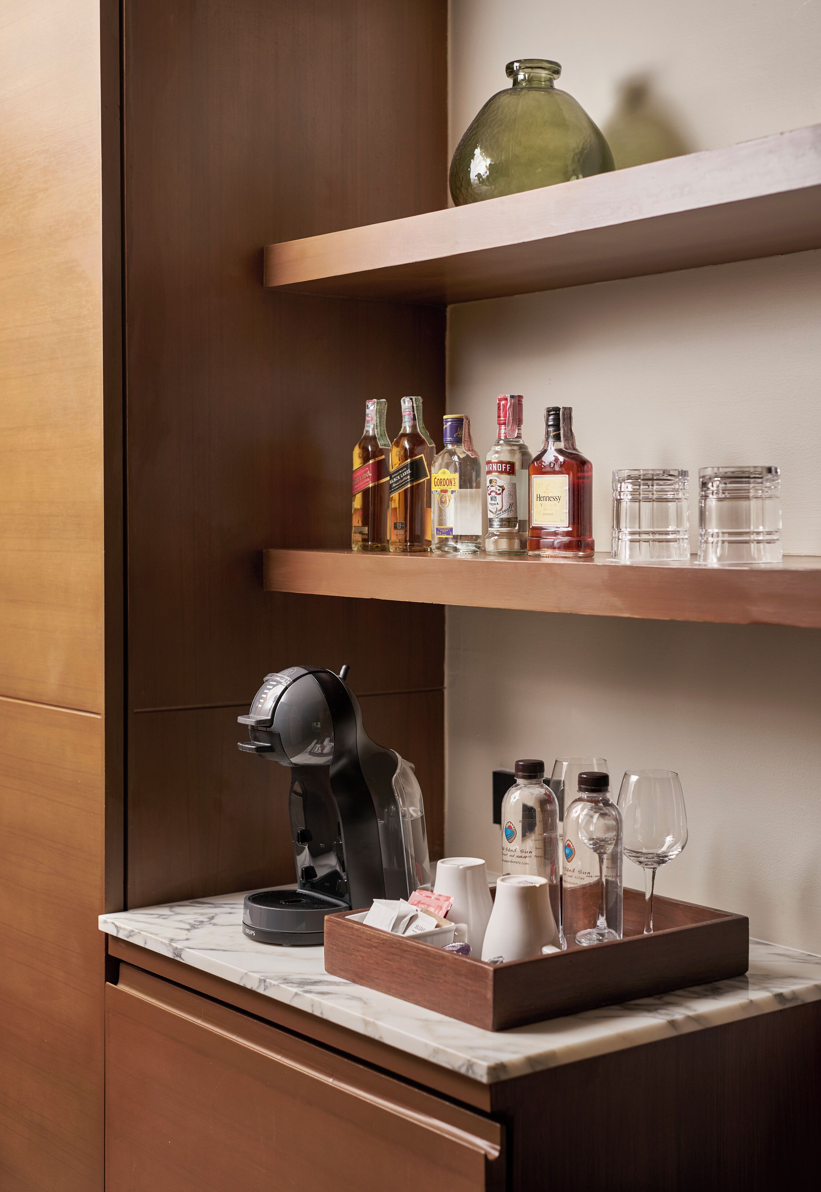A kitchen area with a coffee maker, a tray with glasses and bottles, and shelves with liquor bottles and glasses.