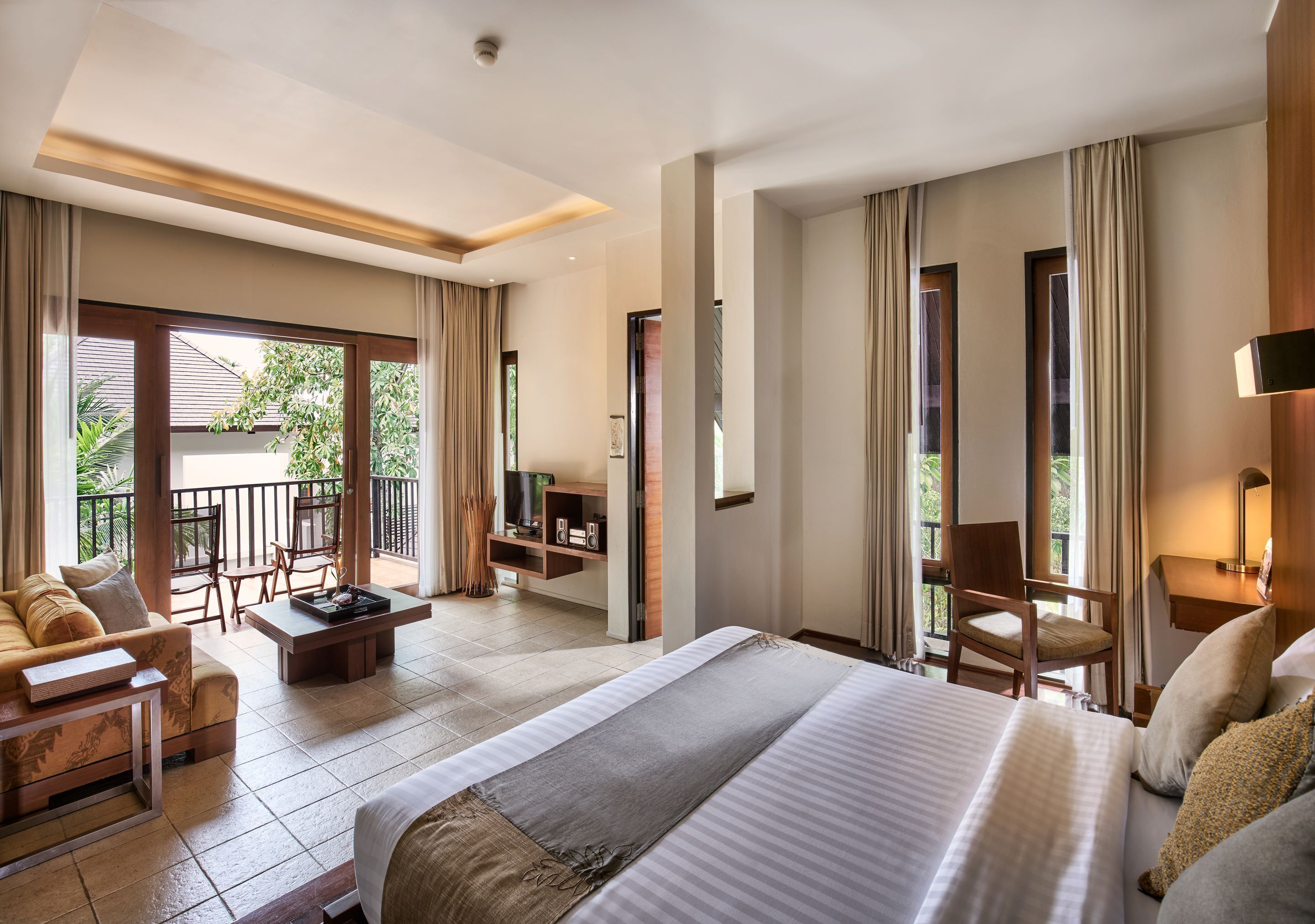 Ein Hotelzimmer mit einem großen Bett, einer Couch, einem Couchtisch, einem Fernseher und einem Balkon mit Blick auf Grünflächen.