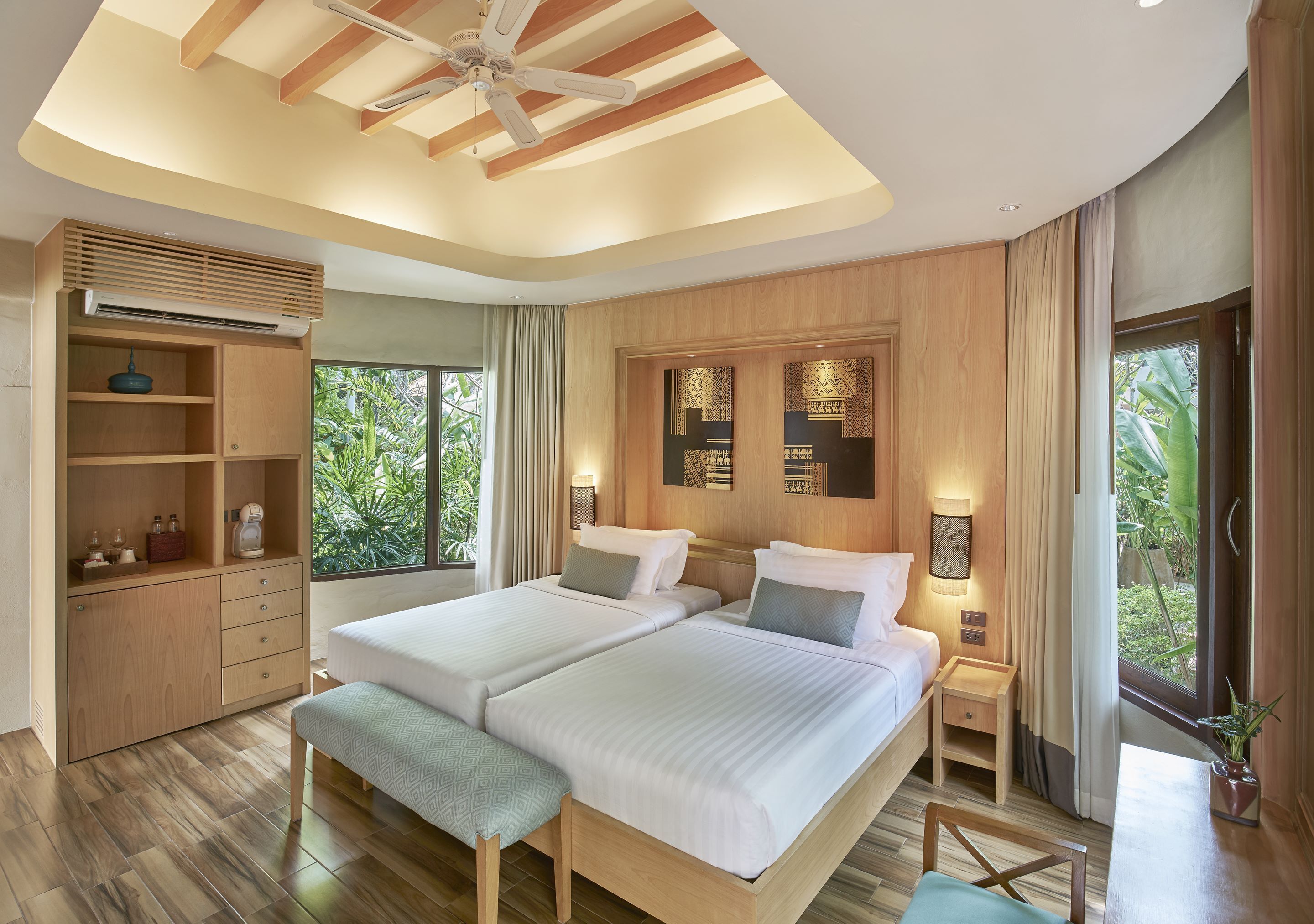 A modern hotel room with two beds, a wooden wardrobe, a ceiling fan, and a large window with a view of greenery.