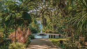 Un sendero serpentea por un jardín exuberante con fuente de agua y un pabellón.