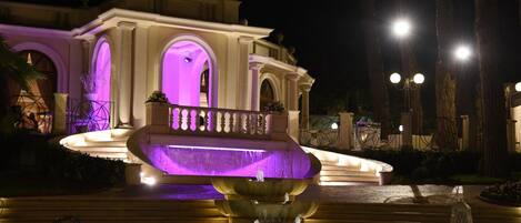 A nighttime view of a grand building with a large fountain in the foreground, illuminated by purple lighting.