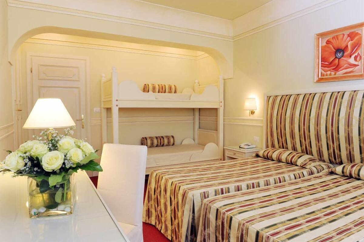 A hotel room with a bunk bed, a single bed, a bedside table with a lamp, a vase of flowers, and a painting on the wall.