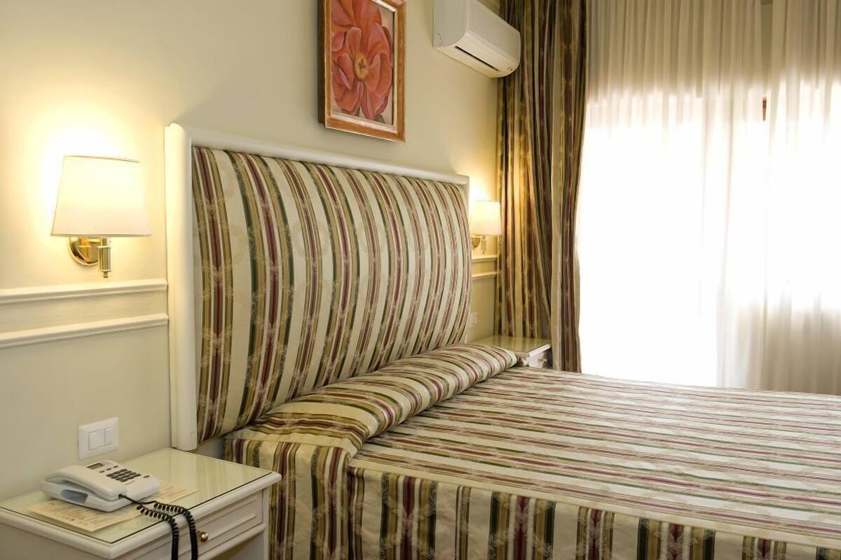 A hotel room with a striped bedspread, a bedside lamp, a telephone, and a painting on the wall.