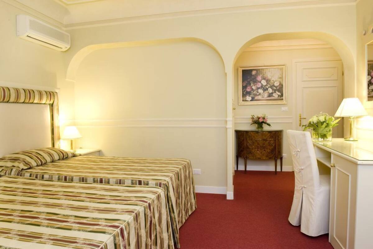 A hotel room with two beds, a desk with a lamp, a chair, and a painting on the wall.