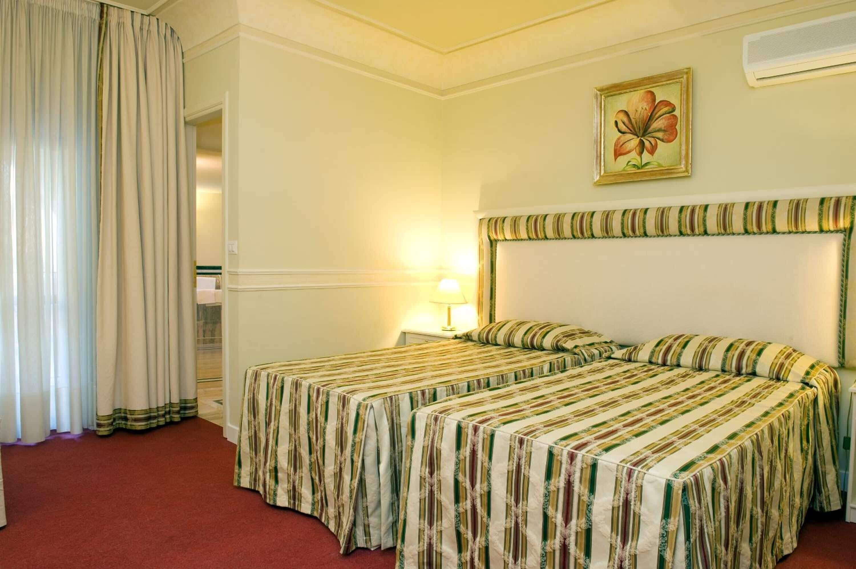 A hotel room with two beds, a nightstand, a lamp, a painting, and a window with curtains.