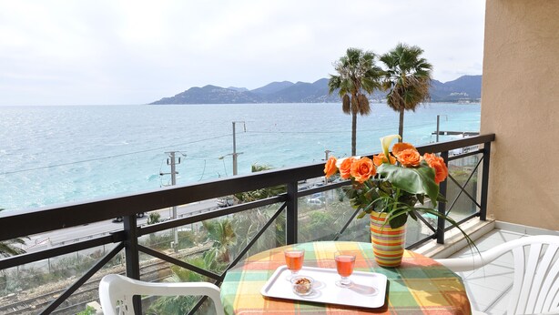 Un balcón con una mesa preparada para dos, un jarrón con flores naranjas y vistas al mar y las montañas.