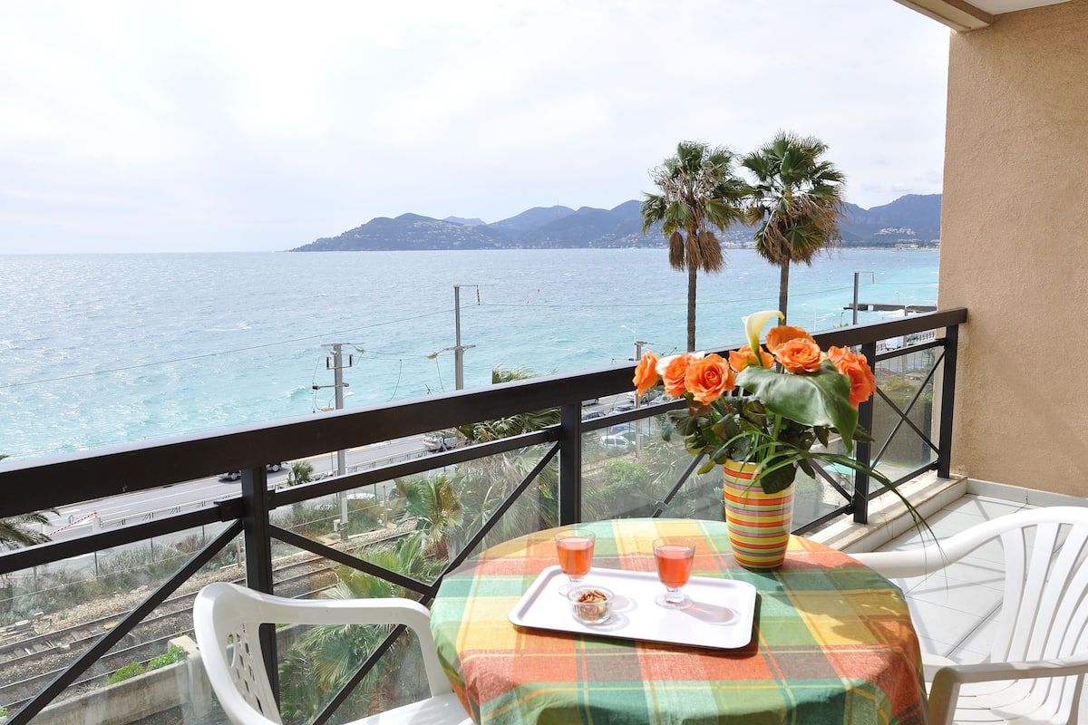 Un balcón con una mesa preparada para dos, un jarrón con flores naranjas y vistas al mar y las montañas.