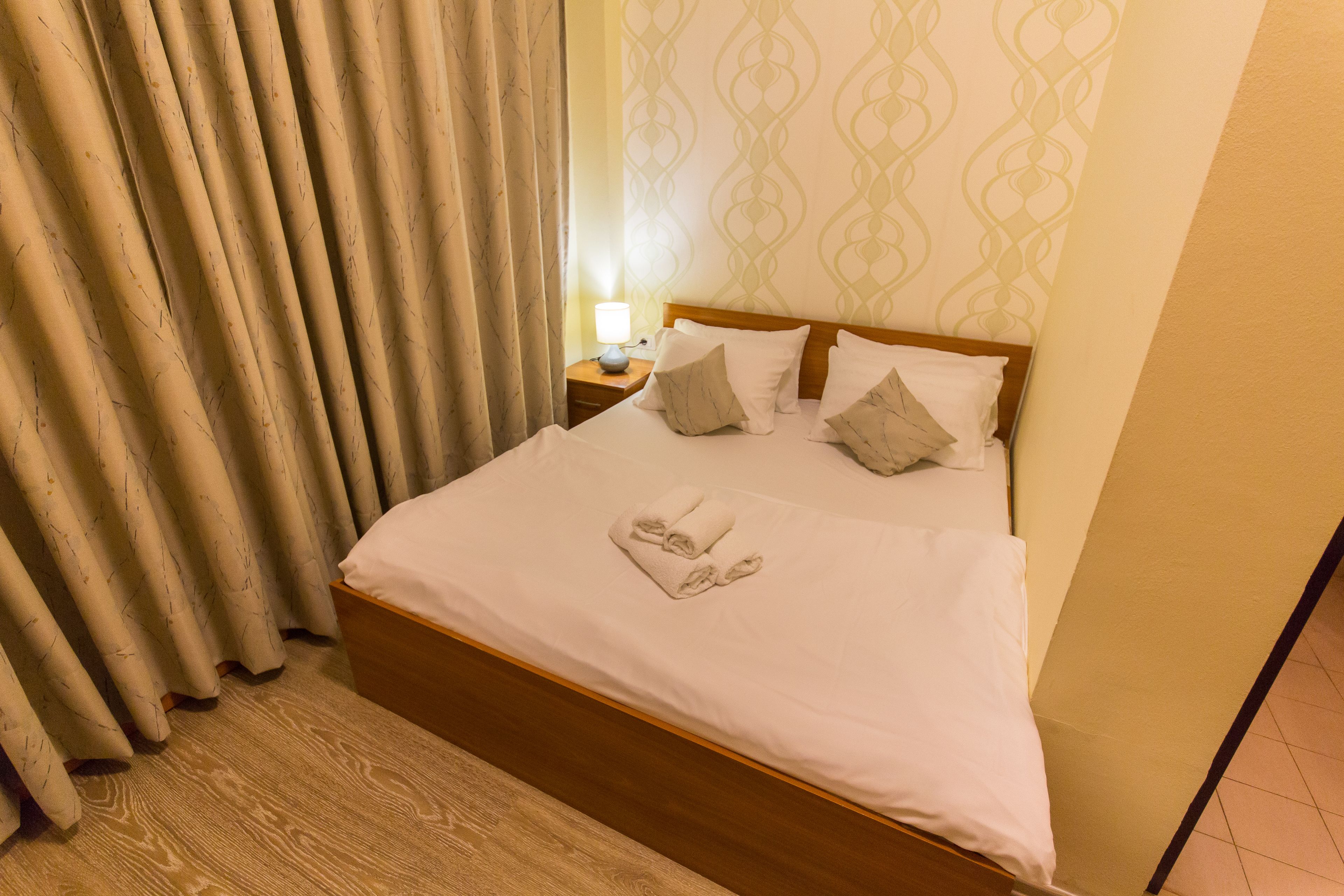 A neatly made bed with pillows and a rolled towel, a wooden headboard, a nightstand with a lamp, and curtains with a leaf pattern.