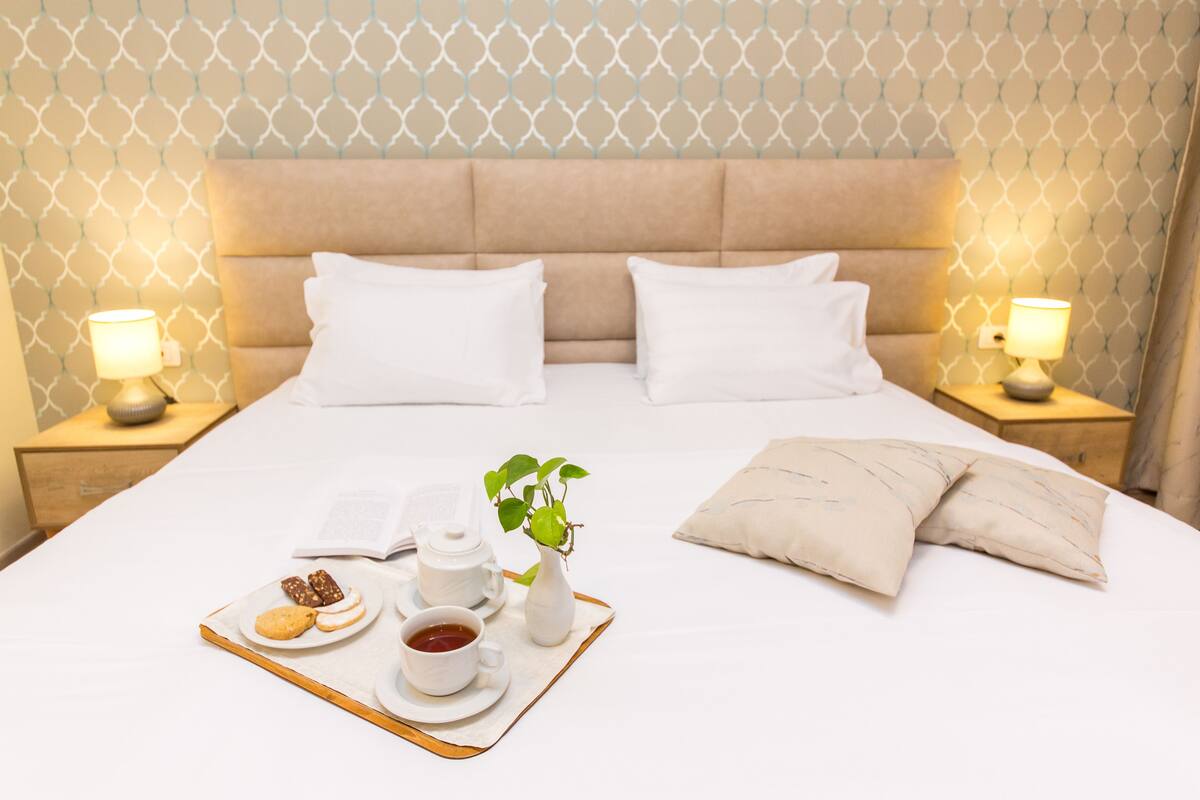 A neatly made bed with white linens, two bedside lamps, a tray with cookies, a teapot, a cup of tea, and a small potted plant.